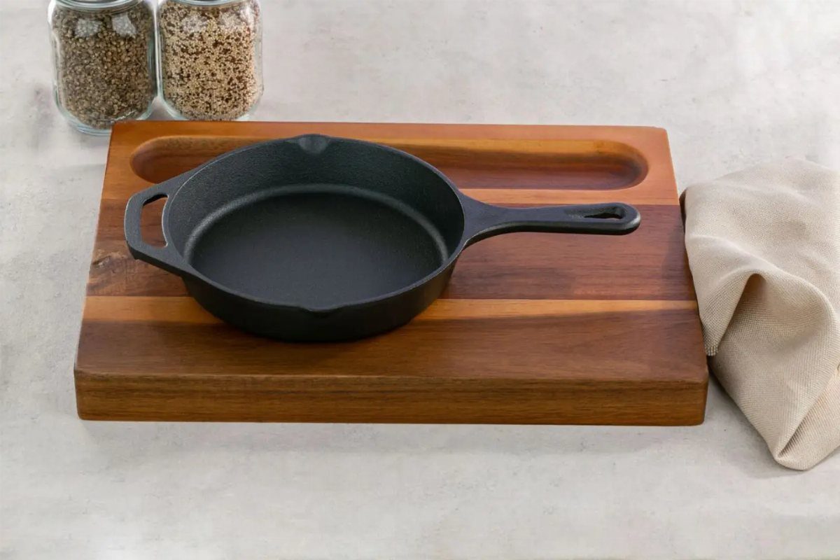 Small Kitchen Essentials: 13 Tools That Actually Matter 9 Black cast-iron skillet on a wooden cutting board with jars of seeds nearby.