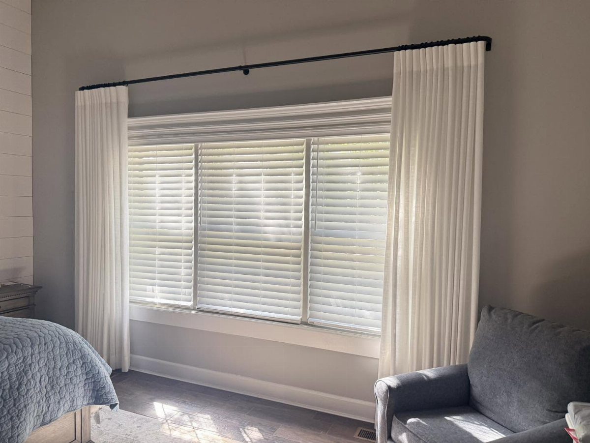 White horizontal blinds paired with white curtains on a black rod in a bedroom.