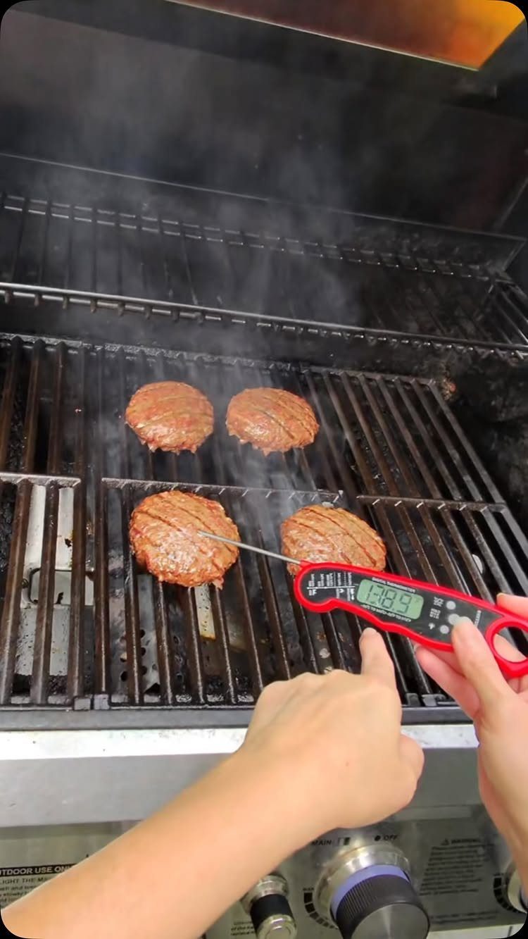 Red digital thermometer probe inserted into a burger patty on a hot grill.