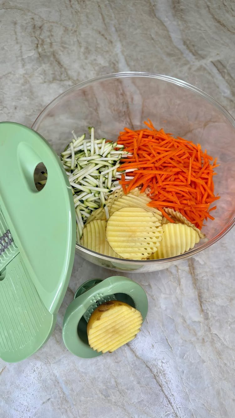 Green mandoline slicer and bowl of sliced carrots, zucchini, and crinkle-cut potatoes.