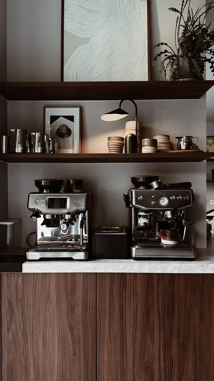 Two espresso machines on a white counter with dark wood floating shelves above.