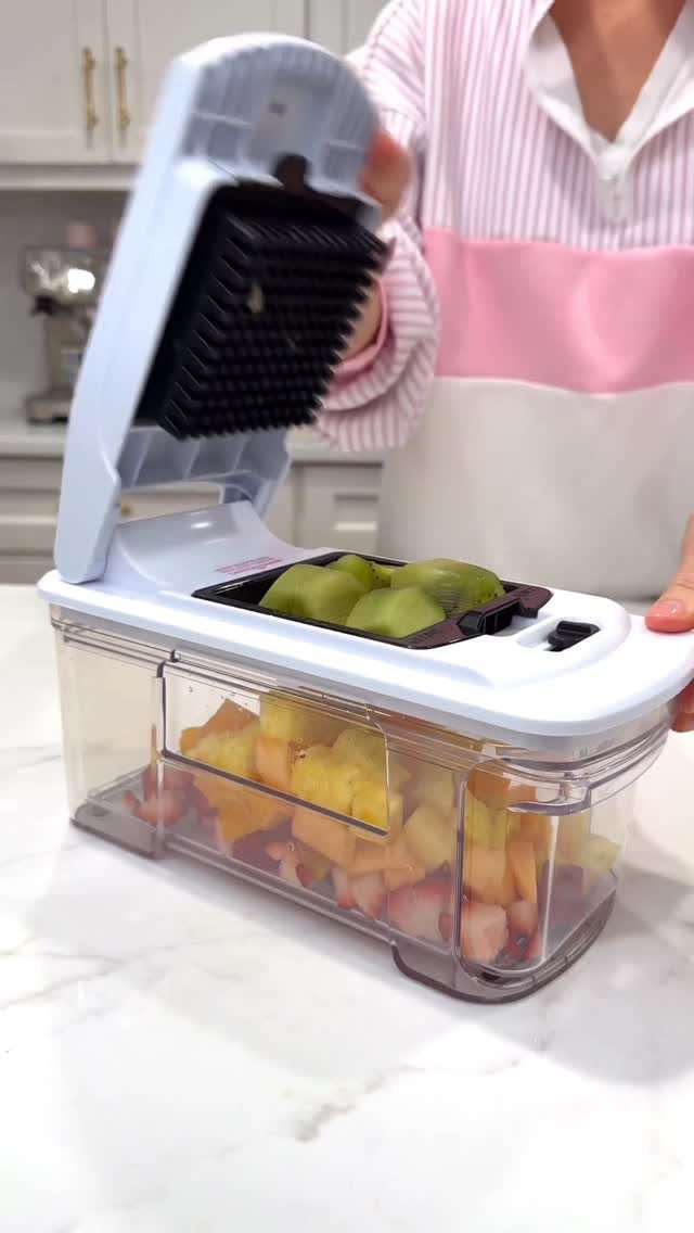 Multi-blade vegetable chopper dicing fruit into a clear plastic container on marble countertop.