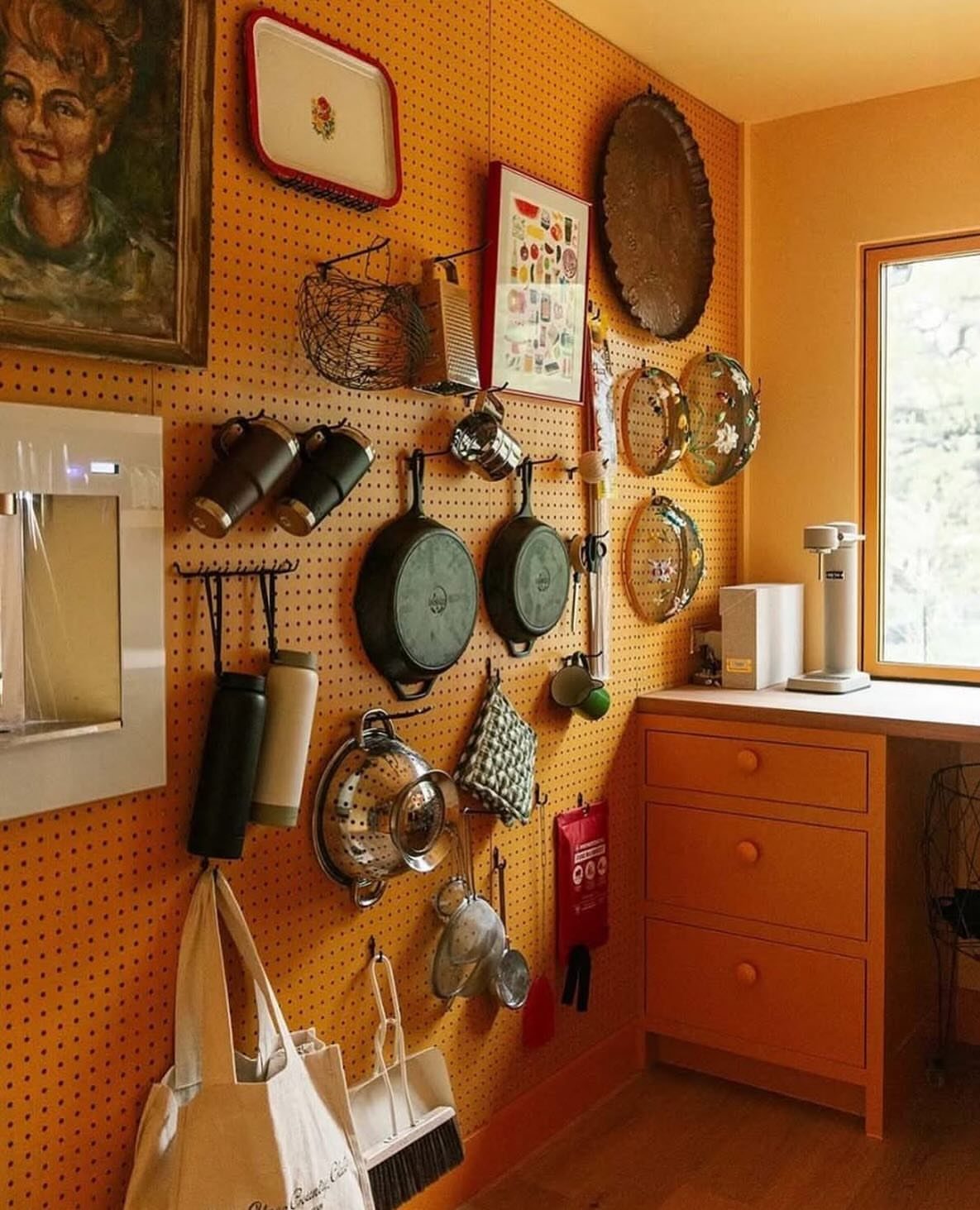 17 Ways to Organize Kitchen Tools (With Photos for Each) 7 Orange pegboard wall holding frying pans, pots, and kitchen tools in a kitchen.