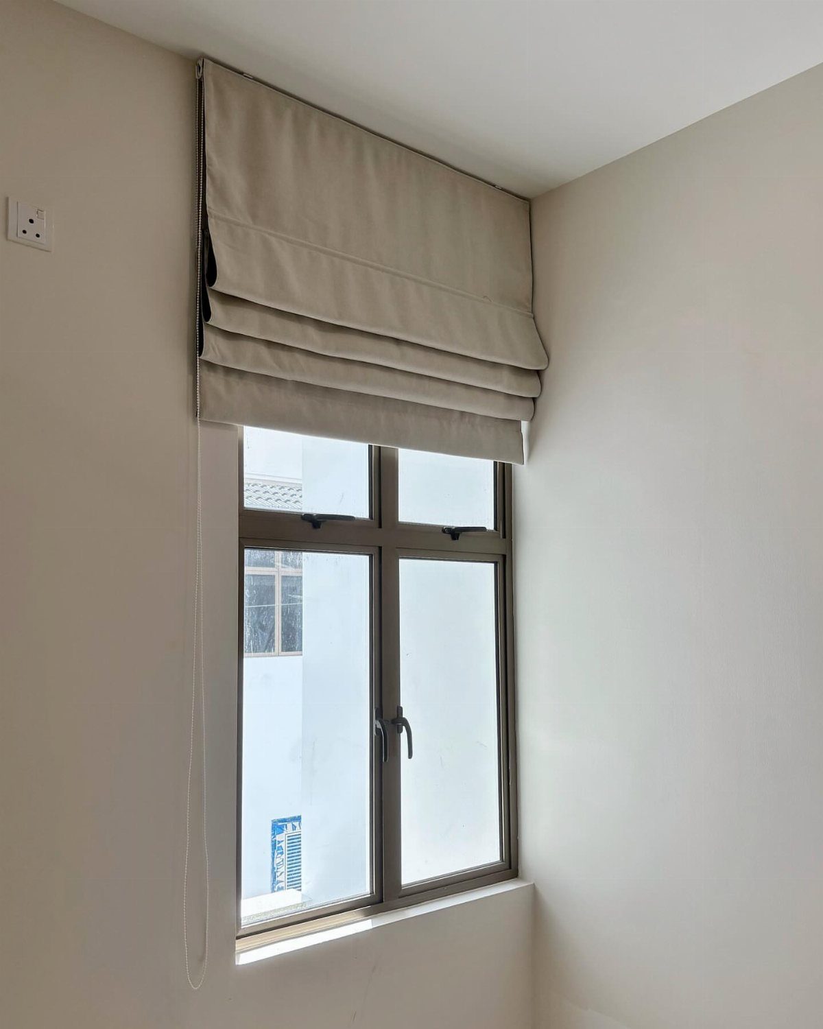 Beige Roman shade with horizontal folds installed on a small white window.