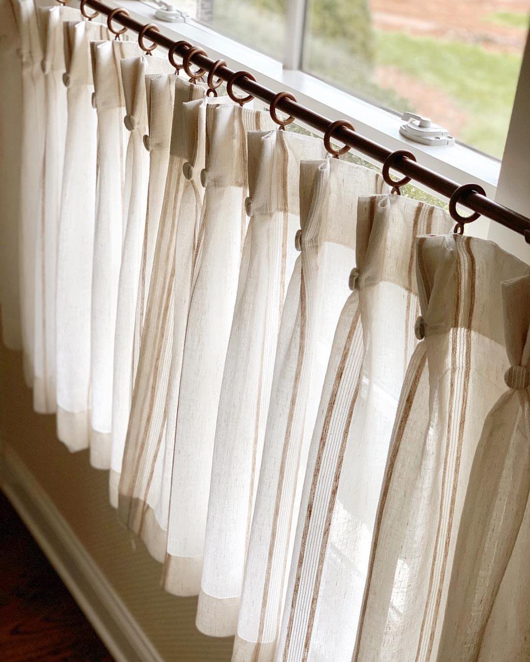 White linen curtains with stripes hanging on a bronze rod by a window.