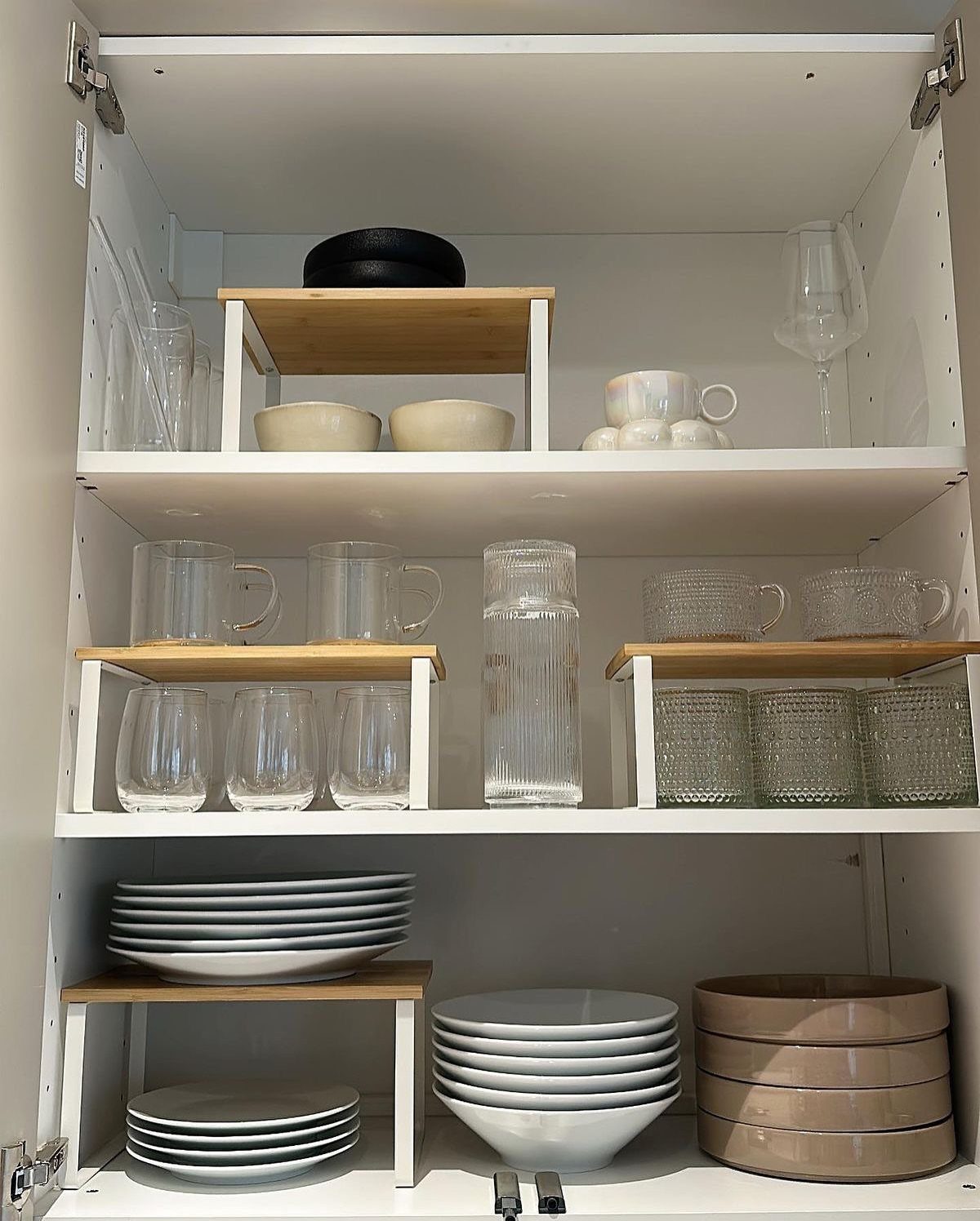 White kitchen cabinet shelves with wooden tiered risers holding glassware and plates.