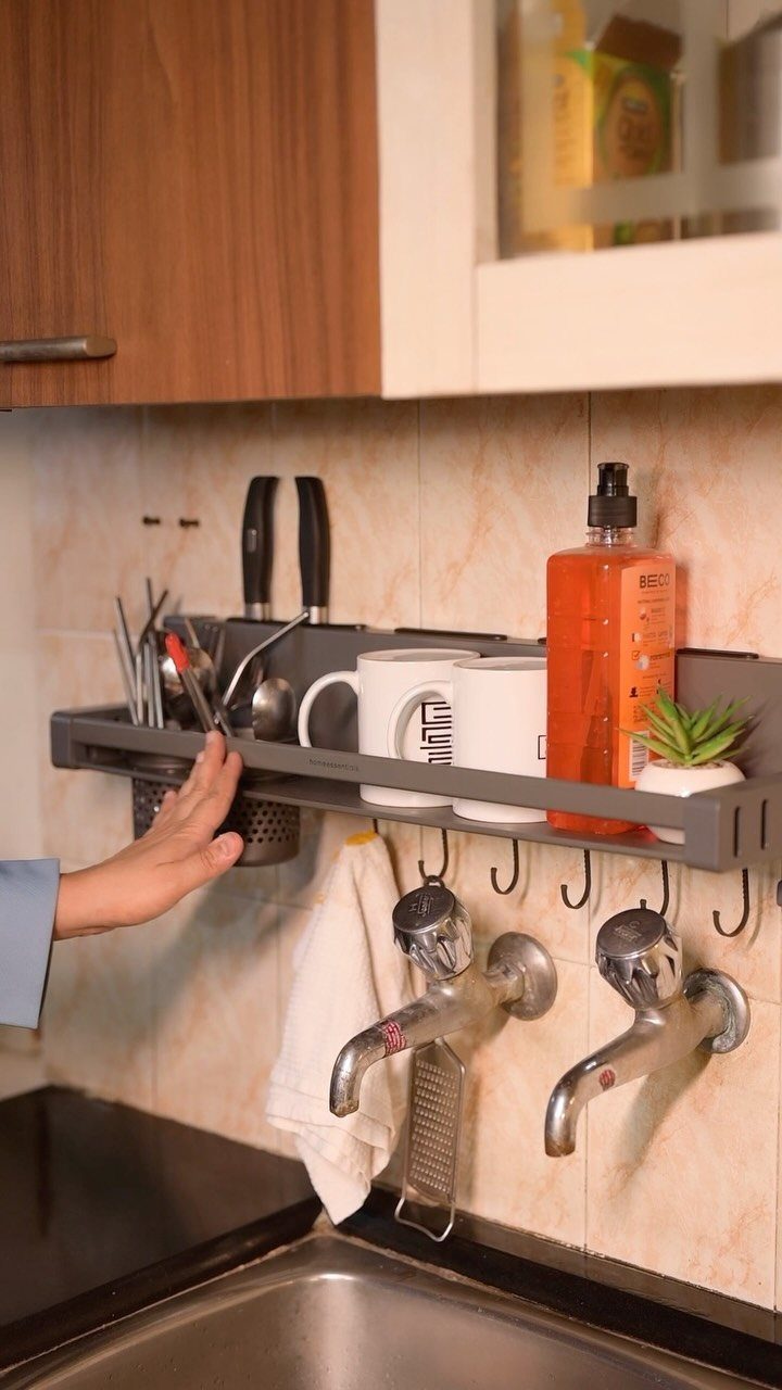Grey wall-mounted rack holding kitchen utensils and mugs above a double faucet sink.