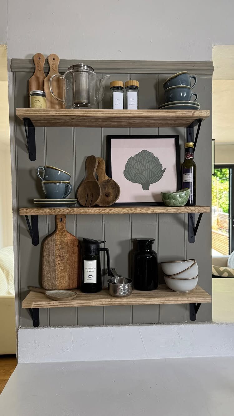 Three wooden wall-mounted shelves holding kitchenware and decor against a grey wall.